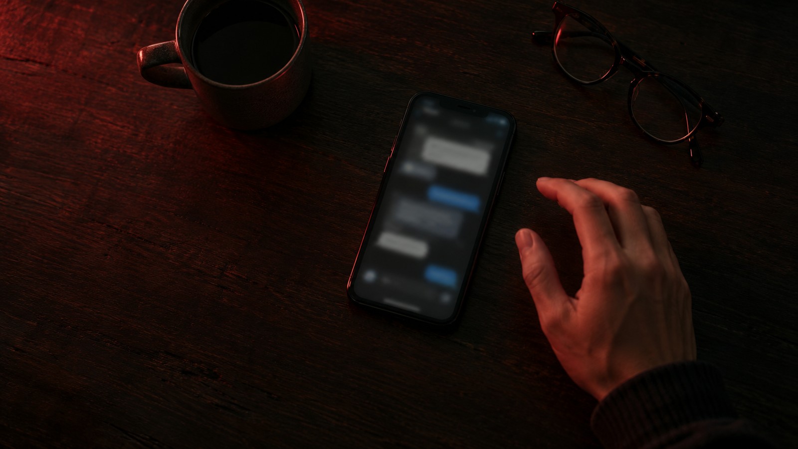 Vista cenital de una mesa de madera oscura con luz roja tenue: una taza, unas gafas y un móvil encendido con una conversación de mensajes ilegible, junto a una mano que se detiene sin llegar a tocar la pantalla